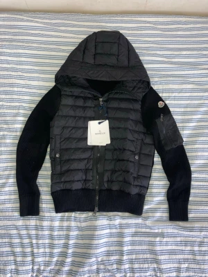 Moncler Cardigan Svart - Snygg svart cardiganjacka från Moncler med quiltad front och huva. Ärmarna är ribbstickade och har en ficka med dragkedja på vänster arm samt Moncler-logga. Jackan har två framfickor med knapp och dragkedja framtill. Perfekt för lager-på-lager och streetstyle. Om du har några frågor om jackan är det bara skriva! Pris kan diskuteras.