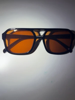 Svarta solglasögon med orange glas - Snygga solglasögon med svart, kantig båge och orangefärgade glas. Modellen har en retroinspirerad design med markerad näsbrygga och breda skalmar. Perfekta för dig som vill sticka ut med en cool accessoar i sommar.