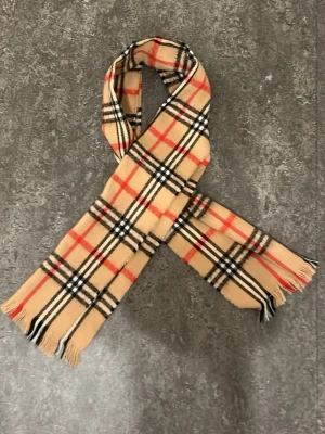 Beige rutig halsduk  - Klassisk beige halsduk med ikoniskt rutmönster i svart, vitt och rött. Halsduken har fransar i ändarna och är tillverkad i mjukt material som känns lyxigt och stilrent. Perfekt accessoar för att lyfta din outfit. Perfekt för kalla väder.