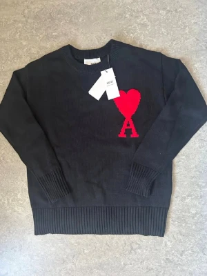 Svart stickad tröja från Ami Paris - Snygg svart stickad tröja från Ami Paris med röd hjärt- och A-detalj på bröstet. Tröjan har rund halsringning, ribbade muddar och nederkant. Perfekt för dig som gillar stilrena plagg med en twist.