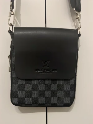 L och V axelväska svart/grå - Snygg axelväska från L och V i svart och grått med klassiskt rutmönster Damier Graphite. Väskan har justerbar axelrem, lock med präglad logga och dragkedjefack på baksidan. Tillverkad i canvas och läderdetaljer.