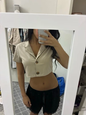 Beige kortärmad crop kofta med knappar - Snygg beige crop kofta med korta ärmar och två stora bruna knappar framtill. Skjortkrage och lätt boxy passform, perfekt för en trendig look. Fint att ha med ett linne men också själv. 