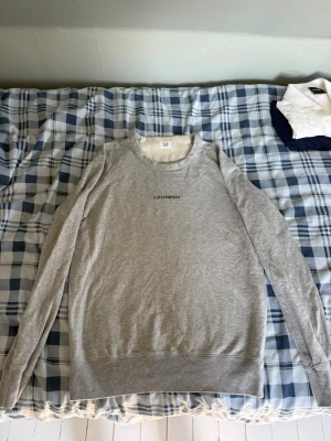Grå sweatshirt från C.P. Company - Snygg grå sweatshirt från C.P. Company med svart logotyp på bröstet. Tröjan har rund halsringning, långa ärmar och ribbade muddar vid ärmslut och nederkant. Tillverkad i mjukt bomullsmaterial som känns skönt mot huden.