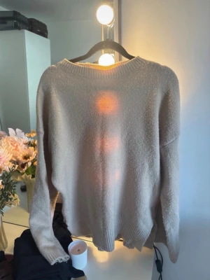 Beige stickad tröja med rund hals - Mysig beige stickad tröja med rund hals och ribbade muddar vid ärmslut och nederkant. Tröjan har en avslappnad passform och är perfekt för lager på lager. Materialet ser mjukt och fluffigt ut, vilket gör den extra skön att bära.