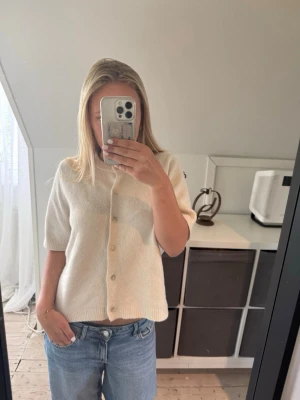 Krämvit kofta med kort ärm från kappahl - Kortärmad kofta i mjuk, krämvit ullmix från Kappahl. Modellen har rund halsringning, ribbade kanter och knäppning framtill med matchande knappar. Perfekt till jeans eller kjol för en clean och enkel look.