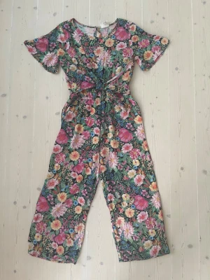 Blommig jumpsuit med knytning - Supersnygg blommig jumpsuit med korta ärmar och knytning i midjan. Färgglatt mönster i rosa, grönt, gult och vitt. Vida ben och rund halsringning. Perfekt för dig som vill sticka ut med en färgstark och bekväm outfit.