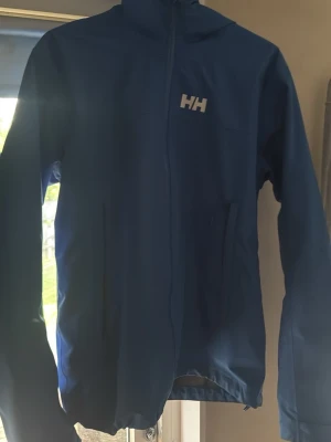 Blå vindjacka från Helly Hansen - Snygg blå vindjacka från Helly Hansen med dragkedja framtill och två sidofickor. Jackan har en hög krage och HH-logga på bröstet. Tillverkad i lätt och slitstarkt syntetmaterial, perfekt för blåsiga dagar. Nypris 1900kr