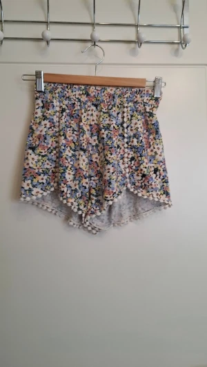 Blommiga shorts med volang från Ardene - Säljer ett par blommiga shorts från Ardene i storlek XS. De har elastisk midja, volangkant och små bollar längs nederkanten. Färgerna är blandade med blått, gult, rosa och vitt. Perfekta för varma dagar och ger en söt och lekfull look.