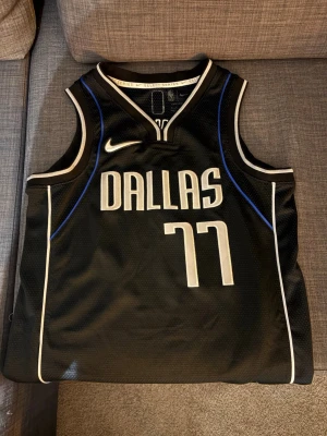 Dallas Mavericks Luka Dončić basketlinne - Snyggt svart basketlinne från Nike med Dallas Mavericks-tryck och nummer 77 för Luka Dončić. Linnet har vita och blå detaljer, broderade loggor och är tillverkat i ventilerande meshmaterial. Perfekt för basketträning eller att bära som streetwear.