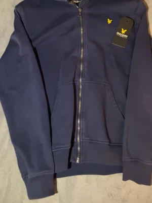Marinblå hoodie från Lyle & Scott - Snygg marinblå hoodie från Lyle & Scott Junior med dragkedja framtill och klassisk gul logga på bröstet. Hoodien har känguruficka och ribbade muddar vid ärmar och nederkant. Tillverkad i mjukt bomullsmaterial för skön känsla.