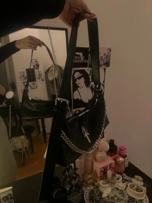 Svart axelväska med kedja och dolde - Cool svart handväska i läderimitation med silverfärgad kedja och ett dolde-emblem på framsidan. Väskan har en justerbar axelrem och dragkedja upptill. 