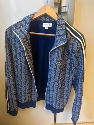 Blå mönstrad ziptröja från Lacoste - Snygg blå ziptröja från Lacoste med vitt geometriskt mönster och klassisk krokodillogga på bröstet. Tröjan har vita ränder längs ärmarna, ribbade muddar och hög krage. Perfekt för en sportig och trendig look. Passar S oxå 