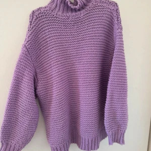 Ljuslila stickad polotröja - Mysig stickad tröja i ljuslila med hög polokrage och breda, avslappnade ärmar. Tröjan har grovstickad struktur och ribbade muddar vid ärmslut och nederkant. Perfekt för lager på lager och ger en trendig oversized look.