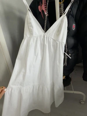 Vit sommarklänning med snörning - Söt vit klänning med tunna axelband och v-ringning. Klänningen har snörning i ryggen och volang nedtill, perfekt för varma dagar. Tillverkad i ett lätt bomullstyg som känns luftigt och bekvämt.