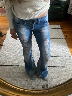 Blå slitna bootcut jeans - Säljer ett par ljusblå bootcut jeans med slitningar och hål på både fram- och baksida. Jeansen har låg midja, klassiska fem fickor och är tillverkade i denim. Perfekta för en avslappnad och trendig look.