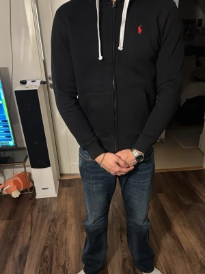 Svart hoodie från Ralph Lauren - Svart hoodie från Ralph Lauren med dragkedja framtill och vit dragsko i huvan. Klassisk röd logga broderad på bröstet. Tillverkad i mjukt material och har två fickor fram. Perfekt för en avslappnad och stilren look. S/M storlek. Fick den i födelsedagspresent Men äger redan en i grå och tycker inte om den svarta så mycke.  Pris gör att diskutera!!!
