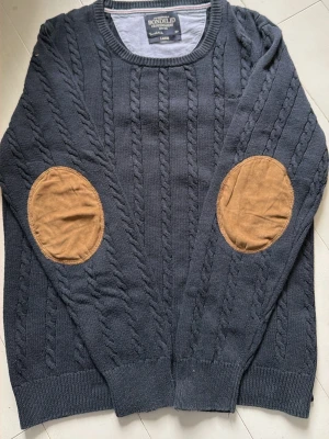 Mörkblå kabelstickad tröja Bondelid - Kabelstickad mörkblå tröja från Bondelid med rund halsringning och ribbade muddar. Tröjan har snygga bruna armbågslappar i mockaimitation som ger en klassisk touch. Hade tänkt använda den men den satt inte som jag hoppades. Den är jätte fräsch och ser knappast använd ut enligt mig. Skriv vid intresse! 