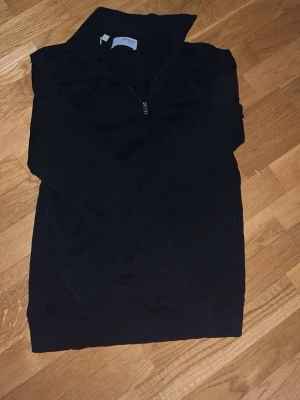 Svart halvzip från Selected Homme - Svart tröja med halv dragkedja från Selected Homme. Klassisk modell med hög krage och ribbade muddar vid ärmslut och nederkant. Perfekt för lager på lager och enkel att matcha med olika outfits.