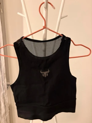 Svart crop top Project Rock meshdetalj - Svart croppad linnetopp från Project Rock med meshdetalj upptill och logga framtill. Toppen är ärmlös och har en sportig look, perfekt för träning eller gym. Snygg och bekväm passform som sitter tight mot kroppen.
