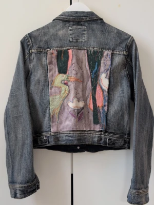 Jeansjacka med broderad ryggpatch - Unik blå jeansjacka med en stor broderad patch på ryggen som föreställer en fågel och blommor i pastellfärger. Klassisk modell med knappar framtill och två bröstfickor. Perfekt för dig som vill sticka ut med en kreativ och personlig stil. Unik och one of a kind. Jeansjackan är peoderad av en konstnär på Österlen!