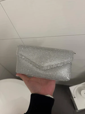 Silverglittrig kuvertväska med strass - Det är en silvrig kuvertväska i silver täckt av  stress med magnetknäppning. Använt en gång 
