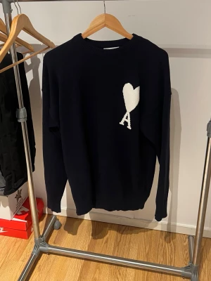 Svart stickad tröja från Ami Paris - Snygg svart stickad tröja från Ami Paris med deras ikoniska vita hjärta och A-logga på bröstet. Tröjan har rund halsringning och långa ärmar. Perfekt för dig som gillar stilrena och trendiga plagg.