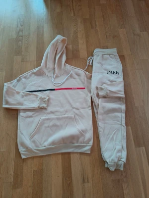 Beige mjukisset med tryck Paris - Säljer ett beige mjukisset med hoodie och byxor. Hoodien har en svart och röd linje med texten 'Paris' framtill, samt känguruficka och dragsko i huvan. Byxorna har resår i midjan, dragsko och texten 'PARIS' på ena benet. Perfekt för en chill look.