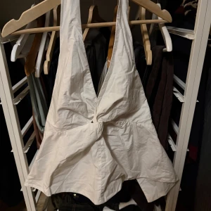 Vit omlottopp från Zara - Säljer en vit omlottopp från Zara med djup v-ringning och breda axelband. Toppen har en twistad detalj framtill och är tillverkad i ett lätt tyg.