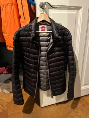 Svart dunjacka från Colmar - Snygg svart dunjacka från Colmar med quiltad design och dragkedja framtill. Jackan har lång ärm och står upp lite i kragen för extra stil. Perfekt för kyliga dagar och enkel att matcha med olika outfits.