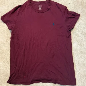 Vinröd t-shirt från Polo Ralph Lauren - Vinröd t-shirt från Polo Ralph Lauren med klassisk rund hals och korta ärmar. Diskret blå logga broderad på bröstet. Perfekt för dig som gillar stilrena basics med en touch av premiumkänsla. Liten fläck längst ner!! Se bild!!