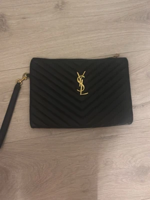 Svart quiltad kuvertväska YSL - Säljer en svart quiltad kuvertväska från YSL med guldigt logotyp-emblem framtill. Väskan har ett elegant mönster, dragkedja upptill och avtagbar handledsrem. Insidan har fack för kort och småsaker. Perfekt för dig som gillar stilrena accessoarer med lyxig känsla.