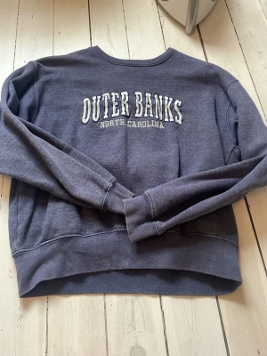 Blå sweatshirt Outer Banks - Mysig blå sweatshirt med broderad text 'OUTER BANKS NORTH CAROLINA' på bröstet. Tröjan har rund halsringning och långa ärmar. Perfekt för en avslappnad stil och passar till jeans eller mjukisbyxor.