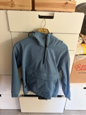 Blå skidjacka från Dope - Snygg blå skidjacka med huva och halv dragkedja framtill. Stor magficka med dragkedja och justerbar nederkant med snören. Jackan har elastiska muddar vid ärmsluten och en minimalistisk logga på bröstet. Perfekt för skidåkning.