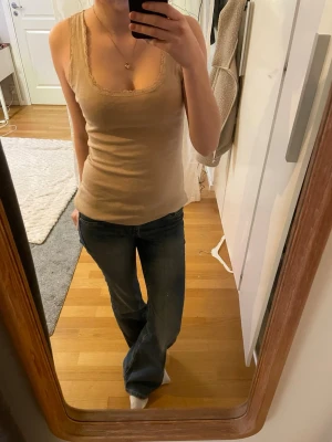 Beige spetslinne med smal passform - Säljer ett beige linne med smal passform och spetsdetaljer längs urringningen och axelbanden. Linnet är ärmlöst och har en mjuk, stretchig känsla som sitter snyggt mot kroppen. Perfekt att styla med jeans eller kjol. Strl M men funkar på mindre 