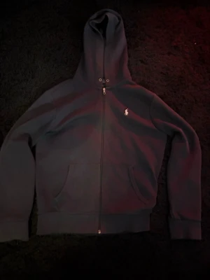 Svart hoodie från Polo Ralph Lauren - Klassisk svart ziphoodie från Polo Ralph Lauren med dragkedja framtill och vit broderad logga på bröstet. Hoodien har huva med snörning och två fickor framtill. Perfekt för en avslappnad och stilren look.