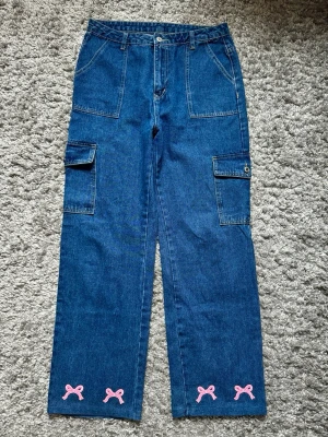Blå cargobyxor med rosa rosetter - Säljer ett par blå cargobyxor i denim med raka ben och stora sidofickor. Nedtill på benen finns söta rosa rosetter broderade som ger en unik touch. Byxorna har hög midja och klassisk cargostil. Perfekta för dig som vill ha något både praktiskt och gulligt.
