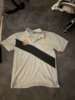 Grå pikétröja från Ralph Lauren - Snygg grå pikétröja från Ralph Lauren med svart diagonal rand över bröstet och broderade detaljer i gult och rött. Klassisk krage och korta ärmar, perfekt för en stilren look. Materialet är mjukt och bekvämt bomullstyg.