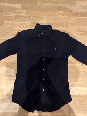 Svart skjorta Ralph Lauren - Säljer en svart skjorta med rött märke från Ralph lauren i mycket bra skick, Inga defekter. Säljer för att den inte passar mig. Tveka inte vid frågor!🤙