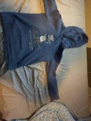 Blå hoodie med björntryck Polo Ralph Lauren - Snygg blå hoodie från Polo Ralph Lauren med ikoniskt björntryck på bröstet. Klassisk huva och långa ärmar. Tillverkad i mjukt material som känns skönt mot huden. Perfekt för dig som gillar streetwear och vill ha en hoodie med lite extra stil.