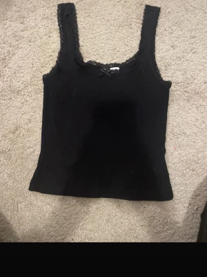 Svart ribbad linnetopp med spets - Snygg svart ribbad linnetopp med spetsdetaljer längs urringningen och axelbanden. Liten rosett framtill för en söt touch. Perfekt att styla med jeans eller kjol för en trendig look.