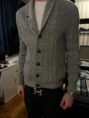 Pier one cardigan - Snygg grå stickad kofta med sjalkrage och svarta knappar framtill. Två fickor fram och bruna mockadetaljer på armbågarna. Ribbstickad struktur och långärmad modell, perfekt för lager-på-lager-stil.