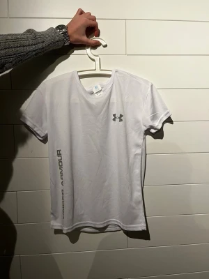 Under Armour T-shirt - Vit sport-t-shirt från Under Armour med rund halsringning och korta ärmar. T-shirten har logga på bröstet och texten 'Under Armour' längs sidan. Tillverkad i ett lätt och ventilerande funktionsmaterial.