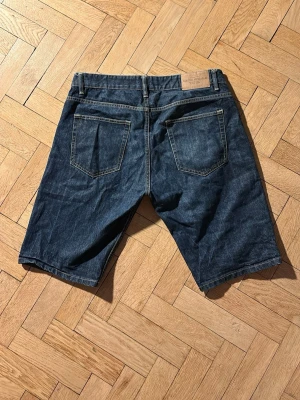 Mörkblå jeansshorts med fem fickor - Säljer ett par klassiska mörkblå jeansshorts med fem fickor och gul kontrastsöm. De har normal passform och är tillverkade i slitstark bomull. Perfekta för sommaren och enkla att matcha med olika stilar.