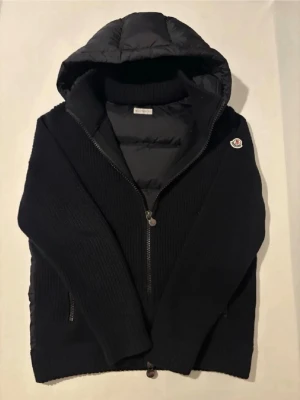 Moncler Cardigan - Svart Cardigan från Moncler med quiltad huva och innerfoder. Jackan har dragkedja framtill, ribbad struktur och Moncler-logga på ärmen. Perfekt för kyliga dagar med sin kombination av stickat material och vadderade detaljer.