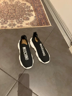 Svarta Moschino skor - Säljer ett par svarta slip-on sneakers från Moschino med vit tjock sula och stor logga på ovansidan. Skorna har en modern, chunky design och är tillverkade i textil med elastisk passform. Perfekta för dig som gillar streetwear och vill sticka ut.