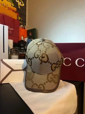 Beige GG-keps från Gucci - Säljer en beige keps från Gucci med det klassiska GG-mönstret över hela kepsen. Kepsen har böjd skärm, justerbart spänne bak och är tillverkad i canvasmaterial med bruna detaljer. Perfekt för dig som vill ha en stilren och lyxig accessoar.