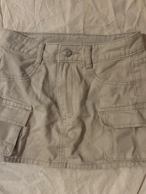 Beige cargo minikjol med fickor - Snygg beige kortkjol i cargo-stil med stora fickor på sidorna och klassisk knappstängning framtill. 