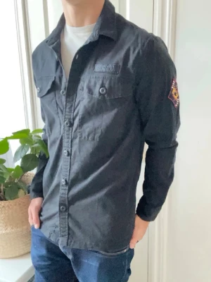 Svart skjorta från Superdry - Säljer en svart skjorta från Superdry med knappar framtill och två bröstfickor. Skjortan har lång ärm och en patch-detalj på ena ärmen. Tillverkad i bomull och har en clean, stilren look som passar till jeans eller chinos.