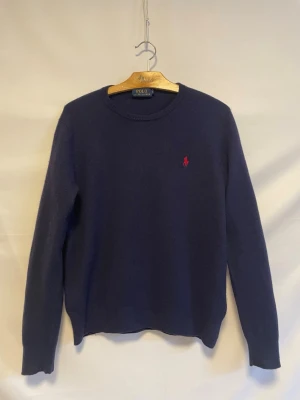 Mörkblå stickad tröja Polo Ralph Lauren - Klassisk mörkblå stickad tröja från Polo Ralph Lauren med rund halsringning och röd broderad logga på bröstet. Tröjan har långa ärmar och är tillverkad i mjukt material som passar perfekt till lager-på-lager-stil.