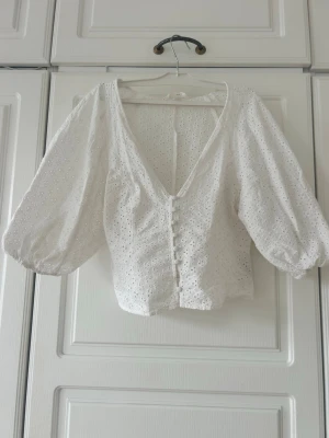 Vit broderad blus med puffärm💖💖 - Söt vit blus med broderade hålmönster och djup v-ringning. Blusen har puffiga trekvartsärmar och knappar framtill. Tillverkad i ett lätt bomullstyg som ger en romantisk och luftig känsla. Perfekt till jeans eller kjol för en trendig look.